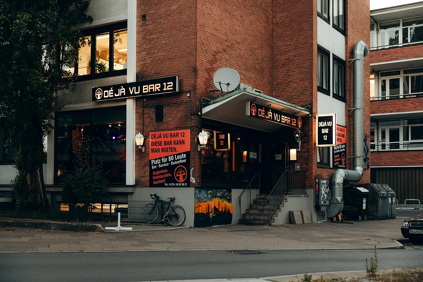 Backsteinfassade der Deja vu Bar 12 mit Leuchtreklame über dem Eingang, Treppe und Plakat „Diese Bar kann man mieten“
