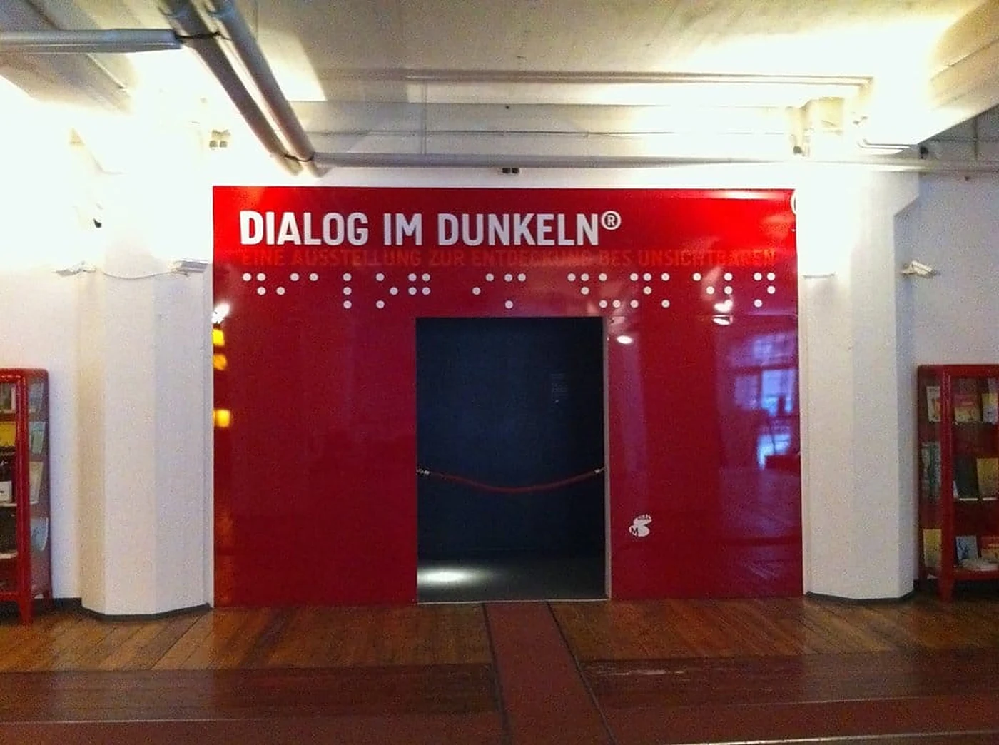Roter Eingang zur Ausstellung Dialog im Dunkeln mit Schriftzug und Brailleschrift, dahinter der dunkle Zugang, davor ein