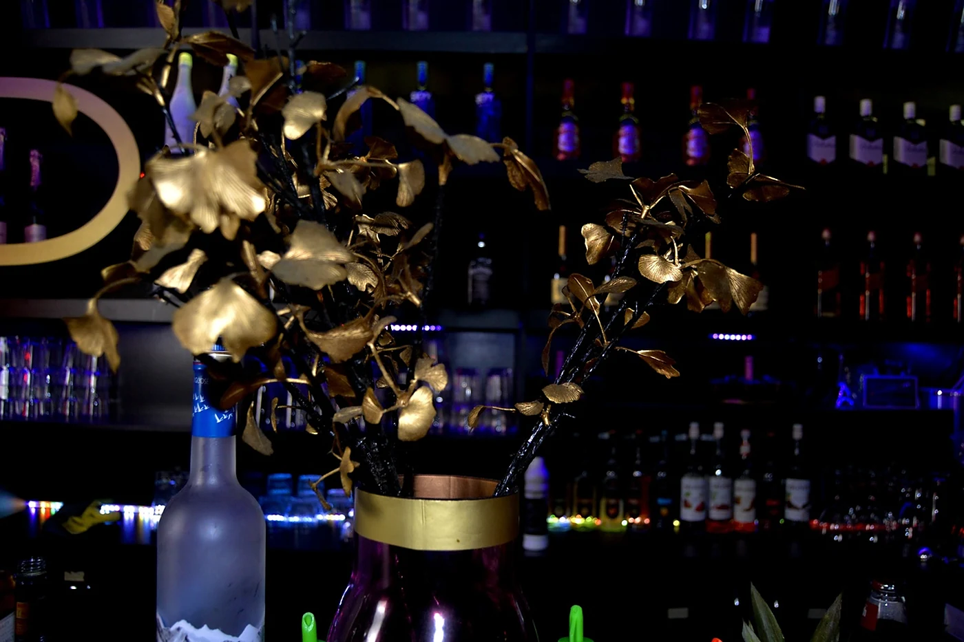 Goldene Blätterdeko in einer Vase vor der blau beleuchteten Flaschenwand der Bar