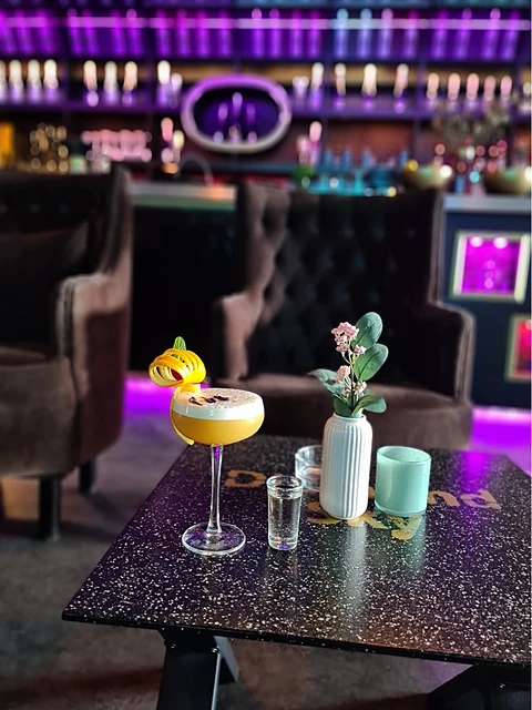 Cocktail mit Physalis im Stielglas auf einem Glitzertisch vor Samtsesseln und der beleuchteten Bar