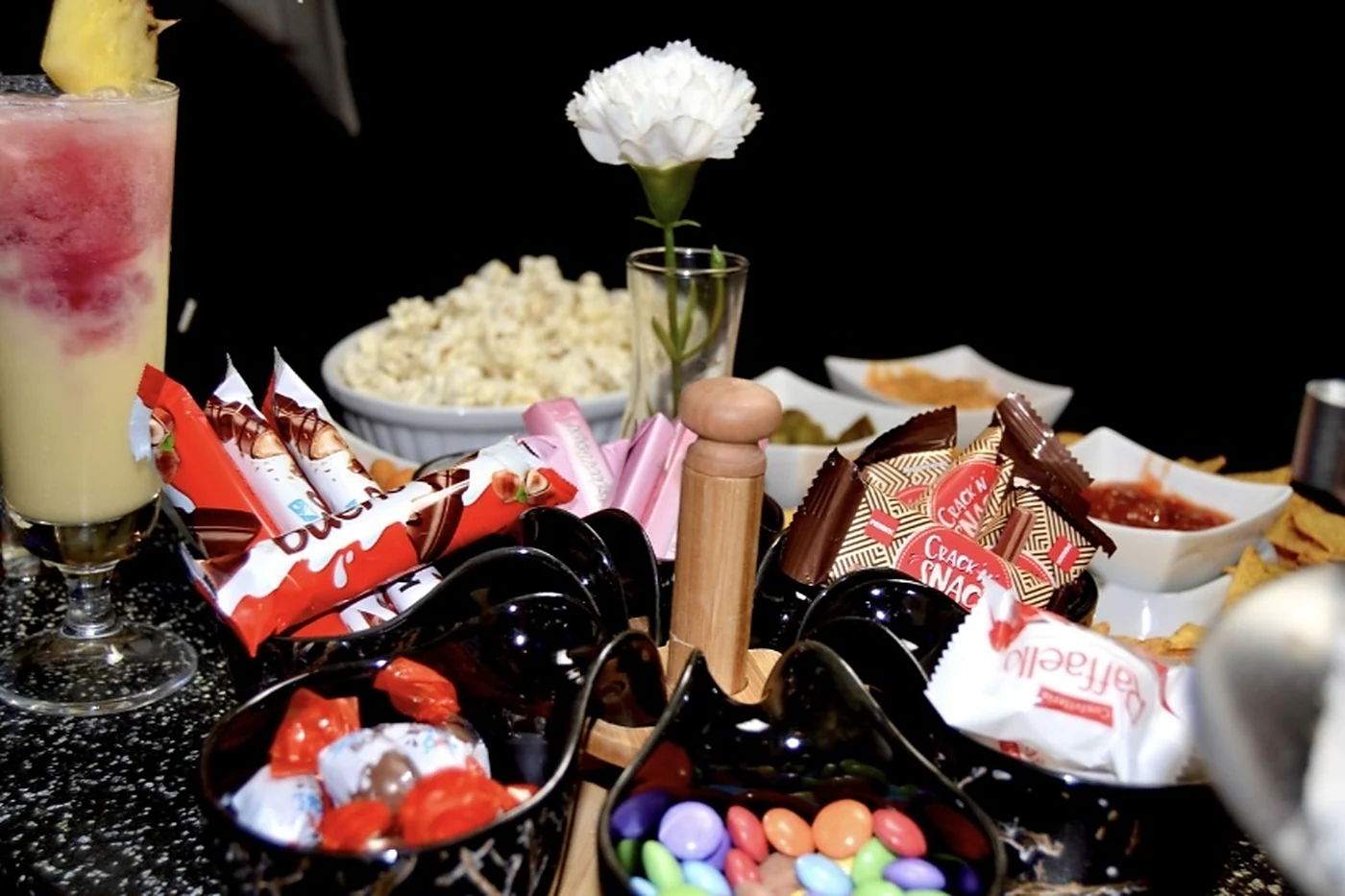Snack-Teller mit Popcorn, Schokoriegeln, Dips und einem rot-gelben Cocktail mit Ananas