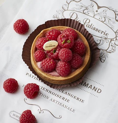 Tartelette mit frischen Himbeeren auf Papier mit dem Logo der Pâtisserie, daneben lose Himbeeren