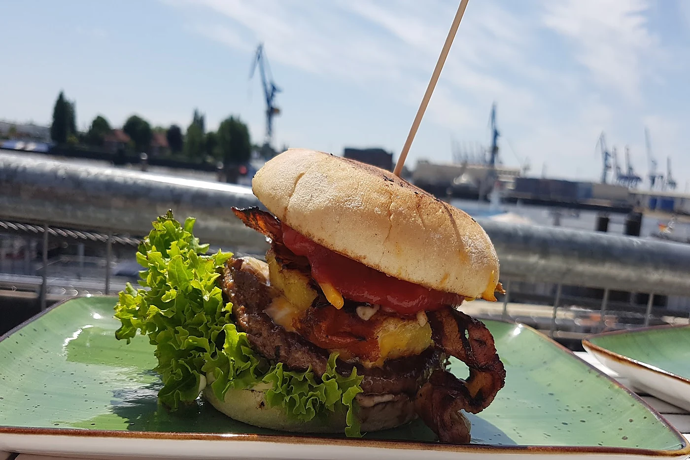Burger mit Speck, Salat und Ketchup auf grünem Teller, dahinter Hafenkrane und Docks