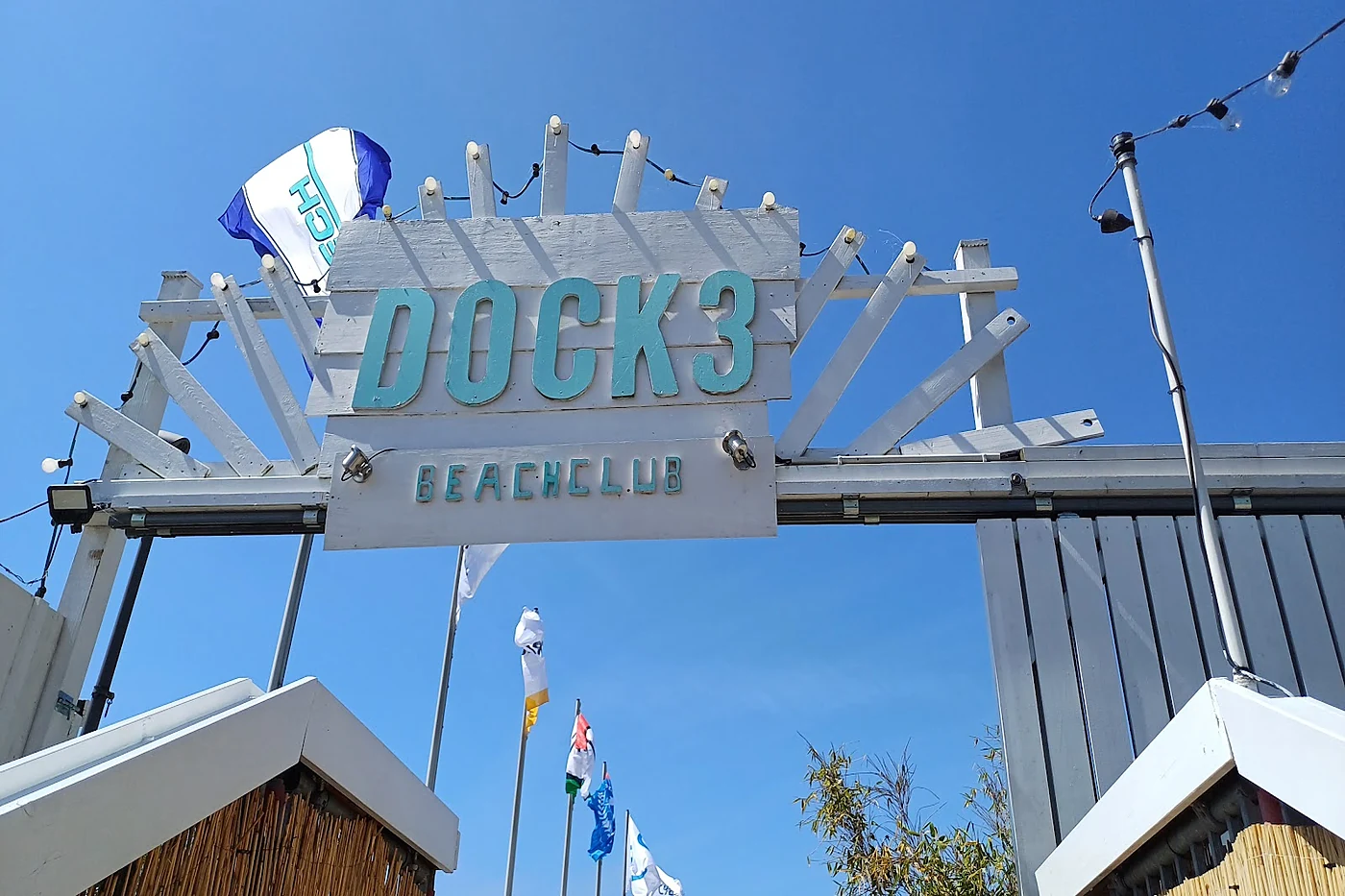 Eingangsschild „Dock 3 Beachclub“ aus weißem Holz vor blauem Himmel mit Fahnen
