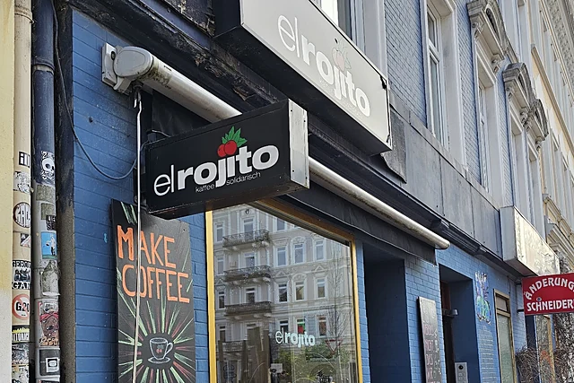 Blaue Backsteinfassade mit el-rojito-Schild und Kreidetafel „Make Coffee Not War“ neben einem großen Schaufenster