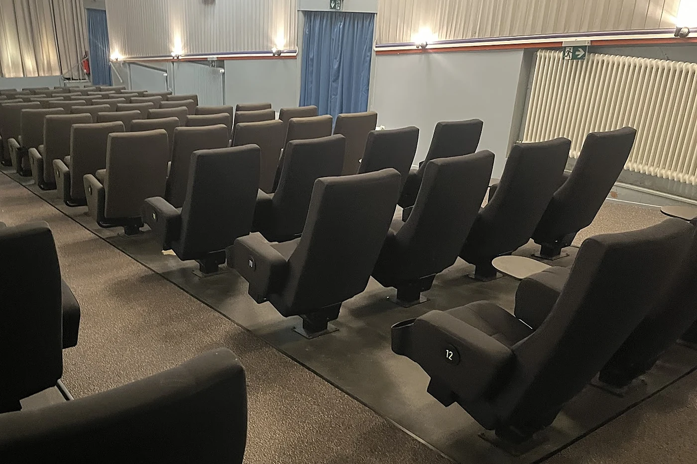 Ansteigende Reihen dunkelgrauer Kinosessel mit Klapptischchen an den Armlehnen in einem hellen Saal