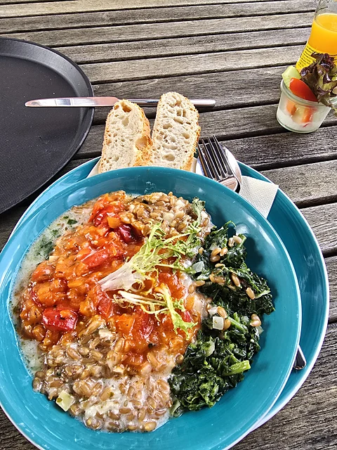 Türkise Schale mit Tomatenragout, Getreide und Grünkohl, daneben Baguettescheiben auf einem schwarzen Teller auf einem H