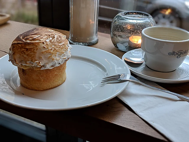 Gebäckstück mit gebrannter Baiserhaube auf einem weißen Teller, daneben Kaffeetasse, Teelicht und Wasserglas auf Holztis