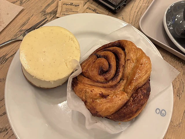 Ein Stück Cheesecake und eine Zimtschnecke auf einem weißen Teller auf Holztisch