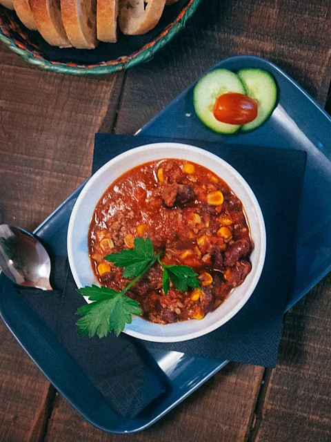 Schüssel Chili mit Mais, Bohnen und Petersilie auf einem blauen Tablett, dahinter ein Brotkorb