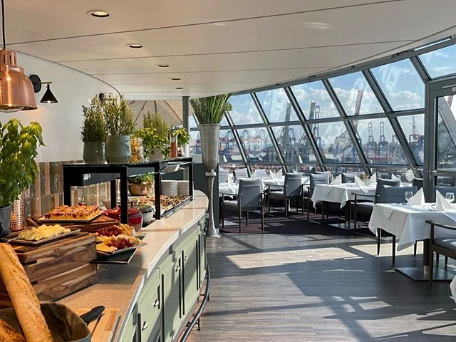 Büfetttheke mit Brot, Gebäck und Obst in der Elbwarte, dahinter gedeckte Tische und Blick auf die Hafenkräne
