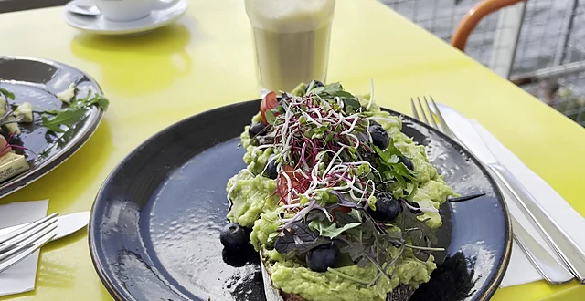 Avocado-Stulle mit Blaubeeren, Sprossen und Tomaten auf schwarzem Teller auf einem gelben Tisch, dahinter ein Latte