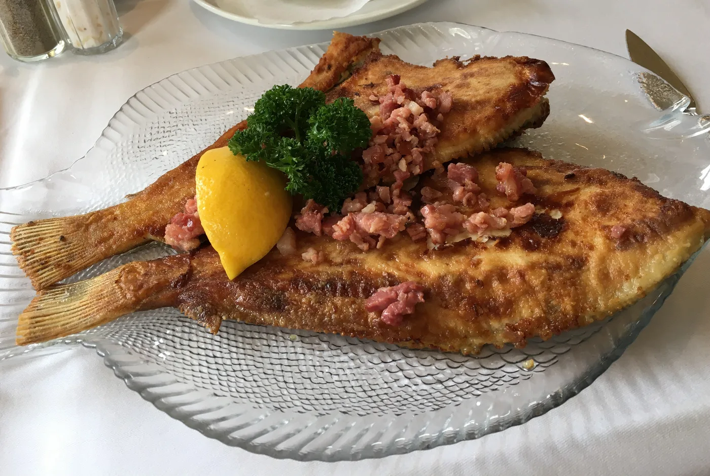 Finkenwerder Scholle mit gebratenen Speckwürfeln, Zitrone und Petersilie auf einem fischförmigen Glasteller