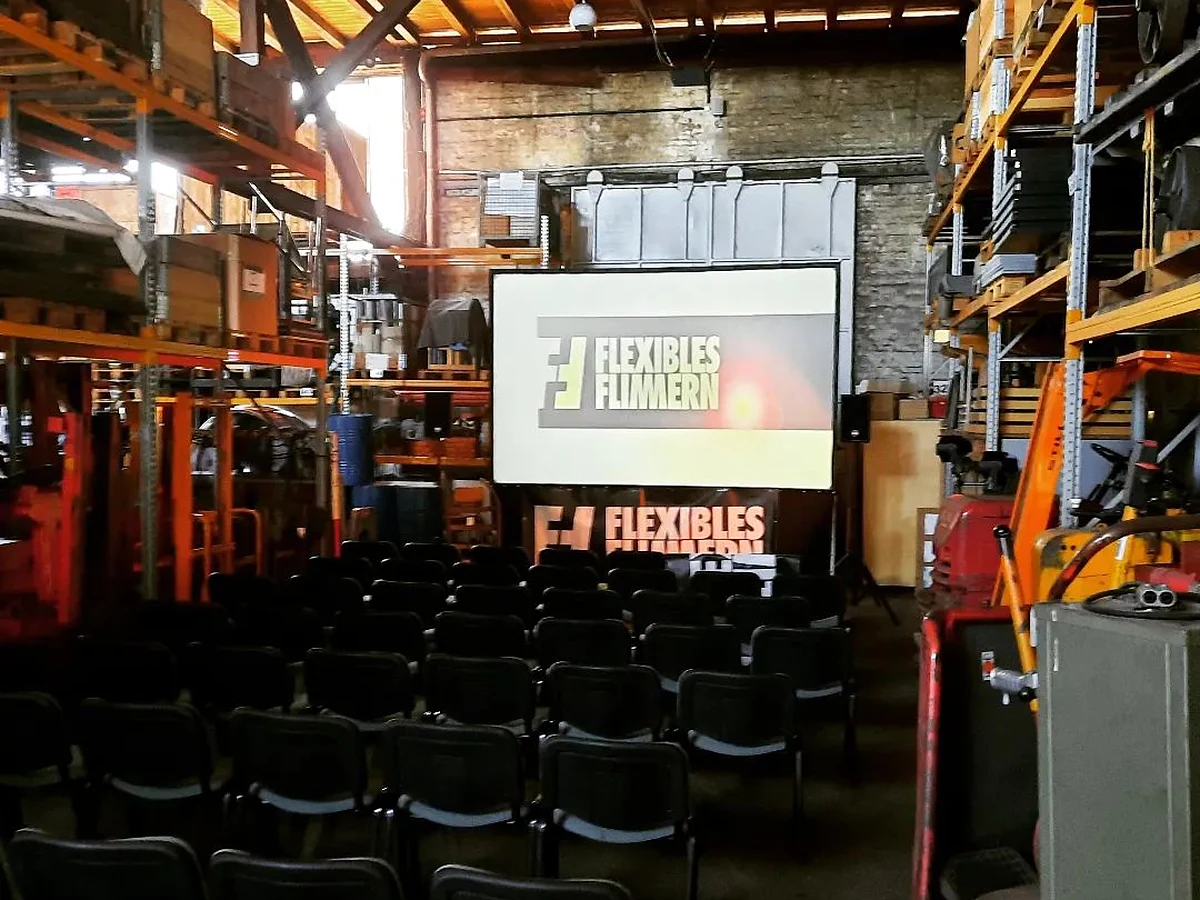 Leinwand mit dem Schriftzug Flexibles Flimmern in einer Halle, davor Stuhlreihen zwischen Regalen und Trägern