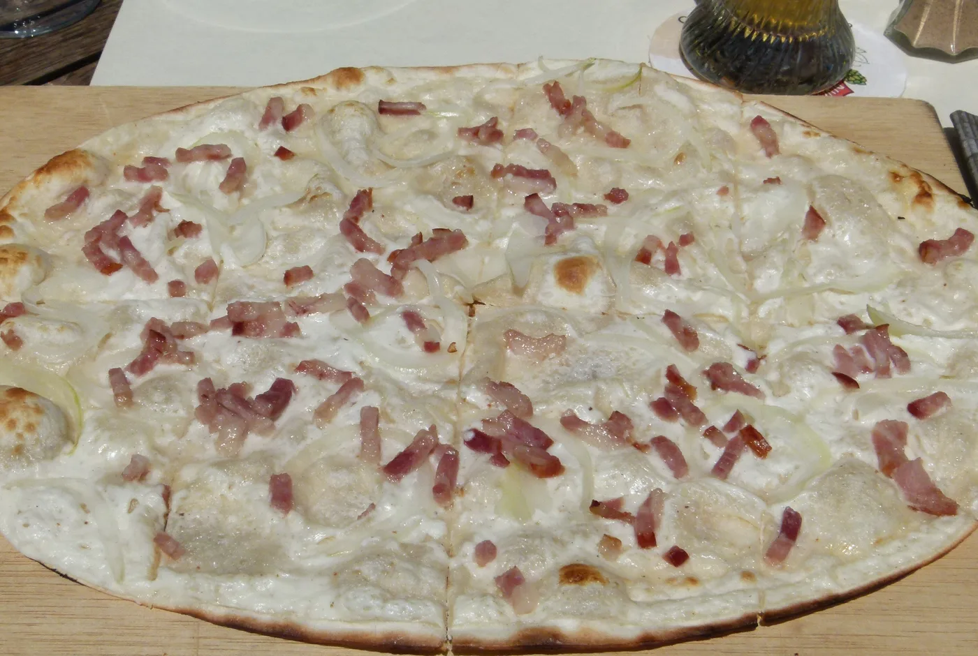 Flammkuchen mit Speckwürfeln, Zwiebelringen und Creme auf einem Holzbrett