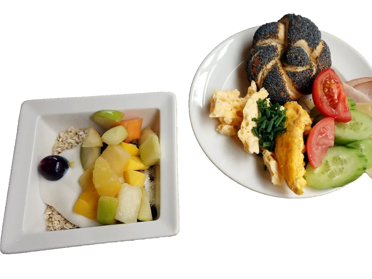 Frühstück mit Müsli und Obst in einer Schale sowie Rührei, Mohnbrötchen, Käse, Schinken, Tomate und Gurke auf einem Teller