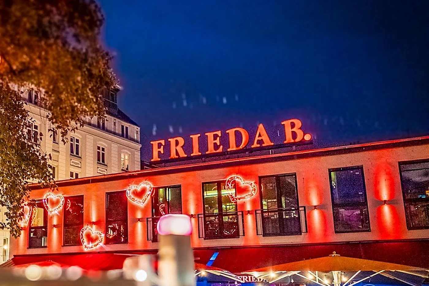 Rot beleuchtete Fassade der Frieda B. am Hans-Albers-Platz mit Leuchtherzen an den Fenstern und dem Schriftzug FRIEDA B. auf dem Dach