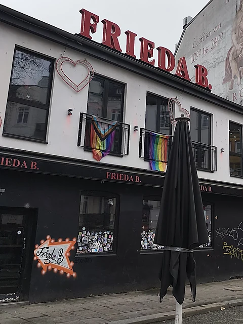 Fassade der Frieda B. in der Friedrichstraße bei Tageslicht mit rotem Schriftzug auf dem Dach, Leuchtherz und Regenbogenfahnen
