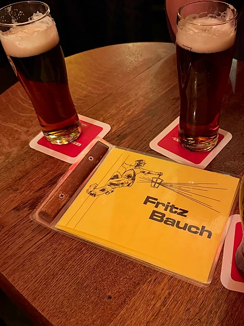 Zwei Gläser dunkles Bier auf roten Deckeln, daneben die gelbe Getränkekarte mit dem Fritz-Bauch-Logo