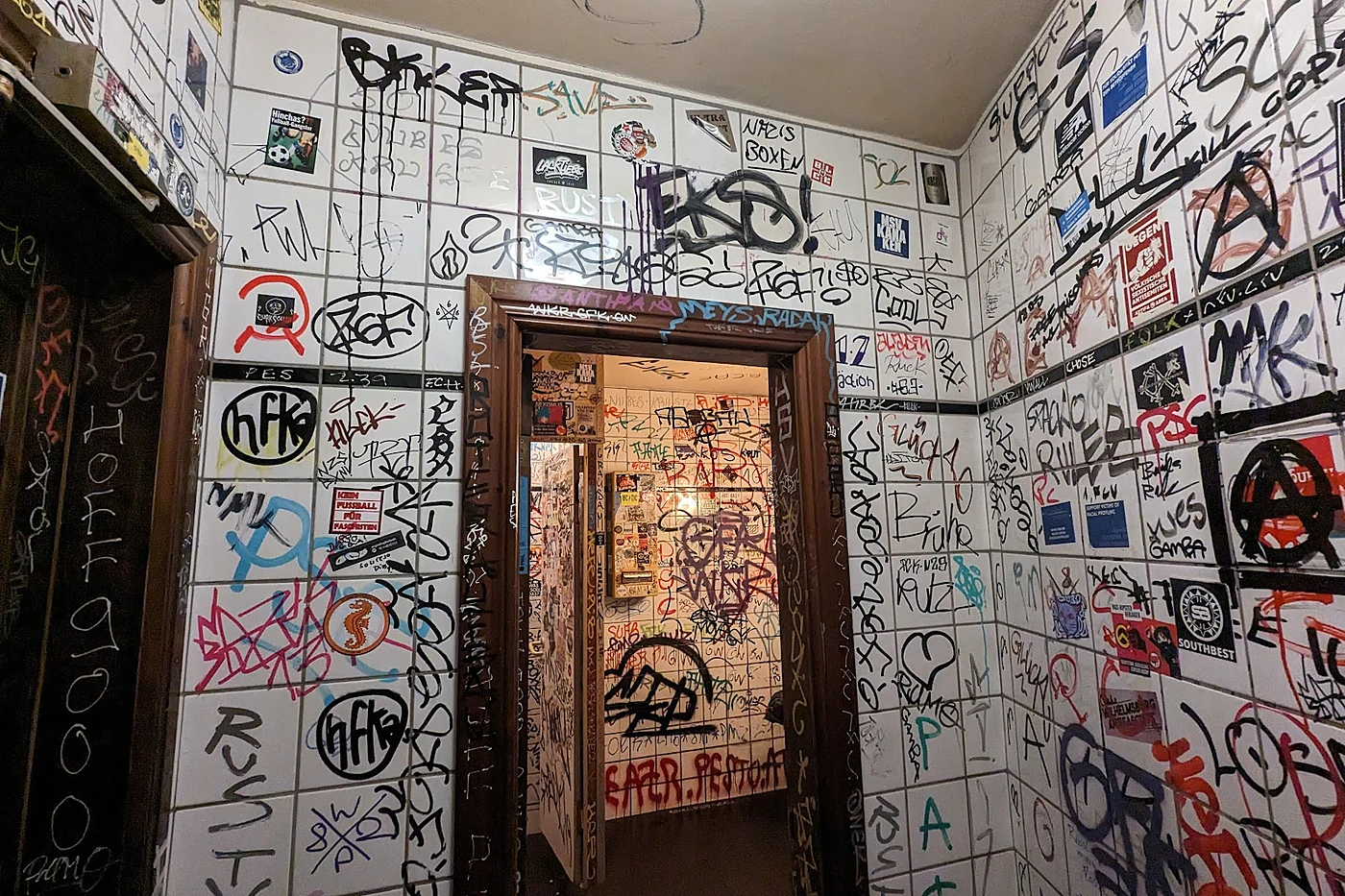 Weiß gekachelter Flur mit dunkler Holztür, jede Fliese mit Stickern, Tags und Kritzeleien bedeckt