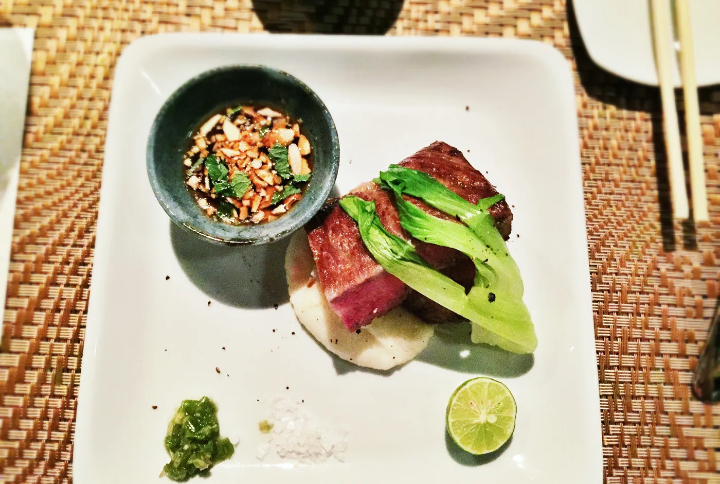 Angebratenes Wagyu-Rind mit Pak Choi auf einem weißen Teller, daneben eine Schale Dip und eine Limette