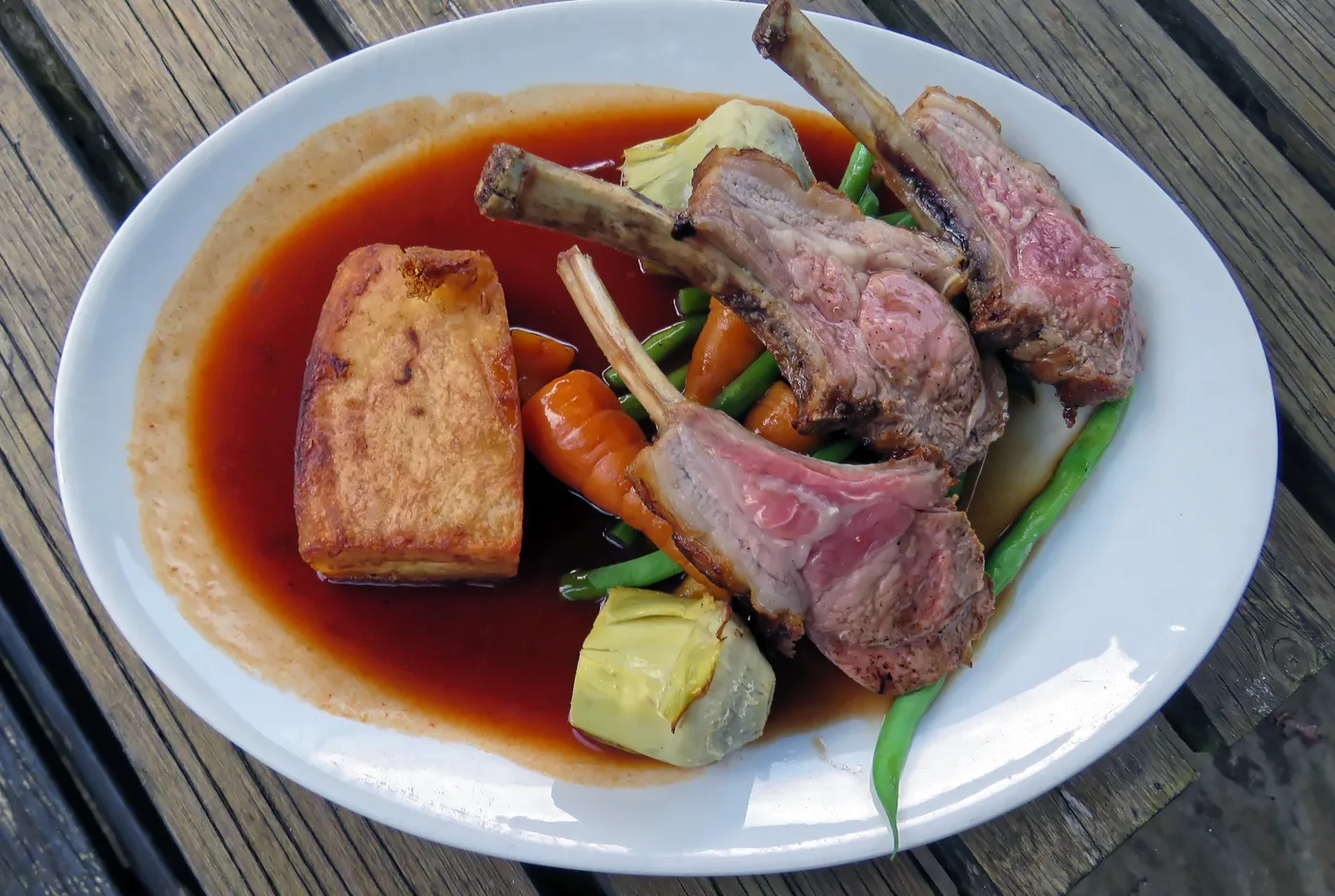 Lammkarree mit Bohnen, Karotten, Artischocken und Kartoffelgratin in einem Jus auf einem weißen Teller
