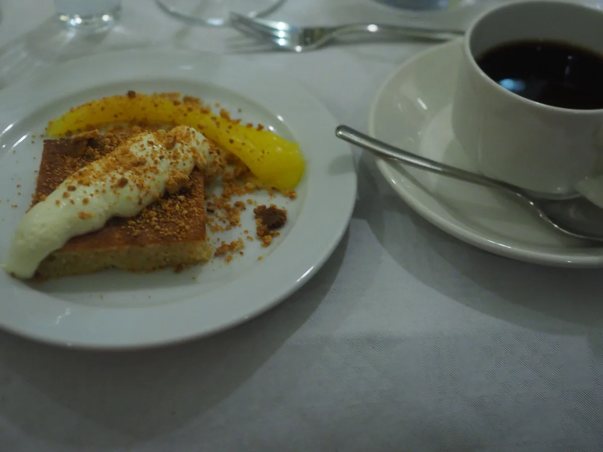 Kuchenstück mit Sahne und Krokant auf einem Teller, daneben eine Tasse Kaffee