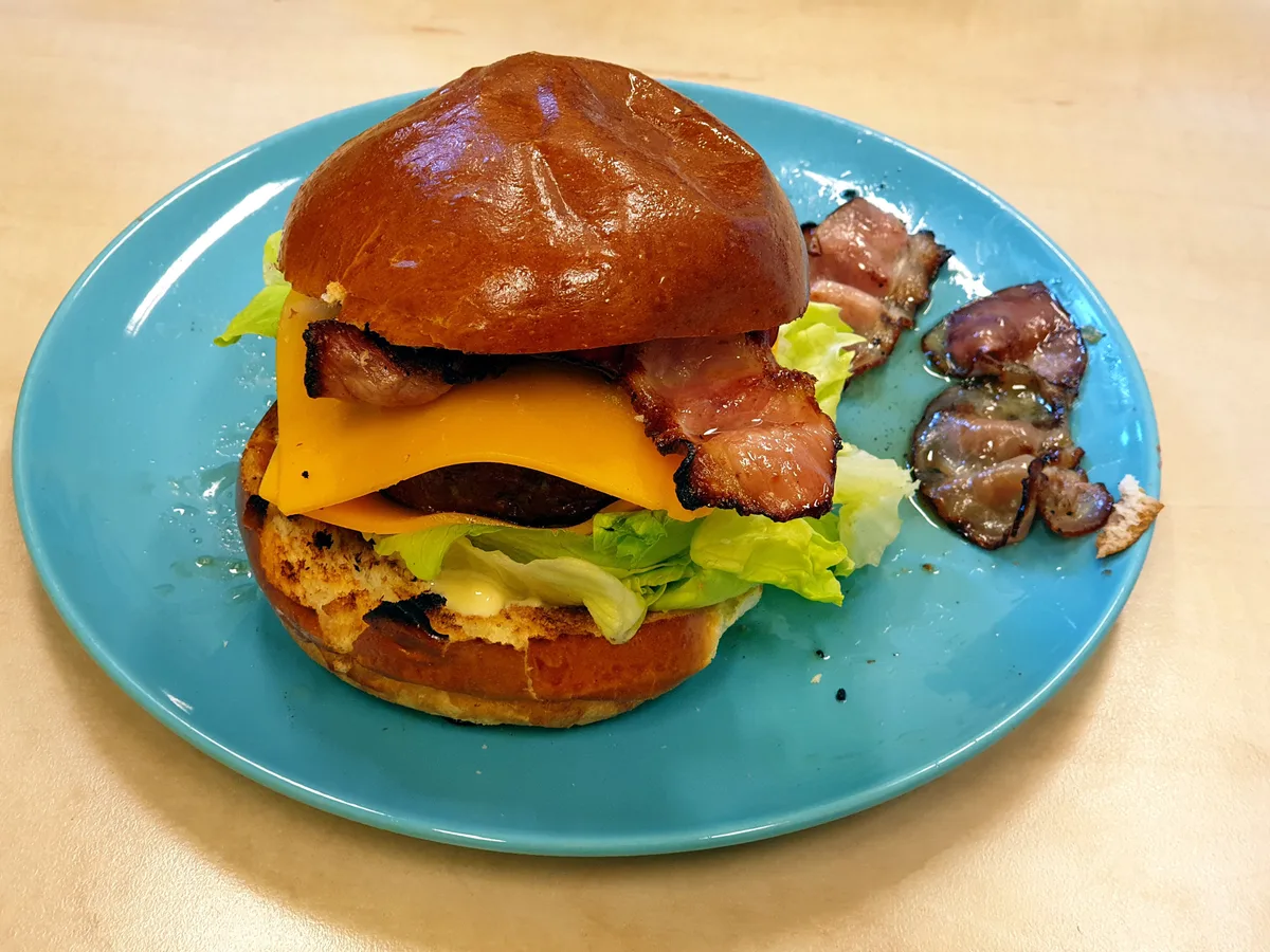 Burger mit Cheddar, Bacon und Salat in einem Briochebrötchen auf einem türkisen Teller