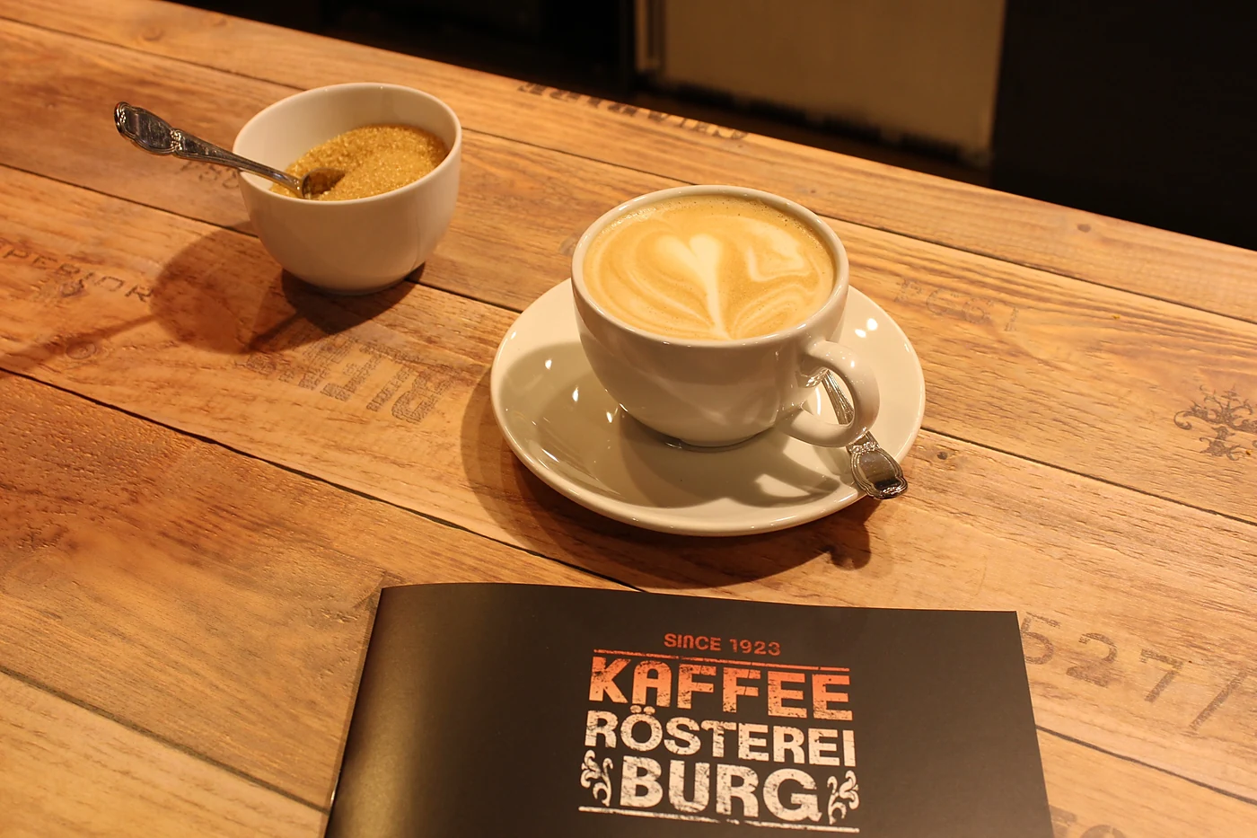 Cappuccino mit Milchschaum-Herz, Schälchen brauner Zucker und eine Karte der Kaffeerösterei Burg auf einem Holztisch im Café