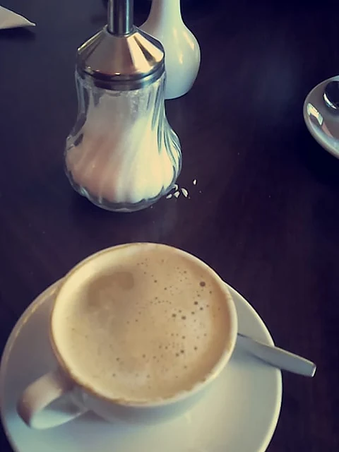Cappuccino in weißer Tasse auf dunklem Tisch, dahinter Zuckerstreuer und Milchkännchen
