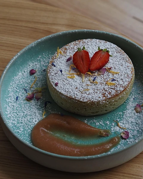 Runder Matcha-Kuchen mit Puderzucker, halbierten Erdbeeren und Blütenblättern auf türkisfarbenem Teller, daneben ein Kle