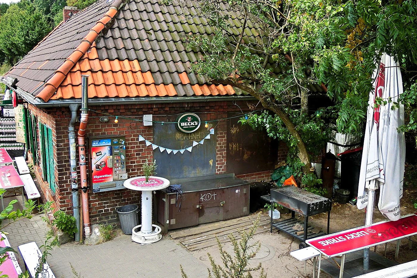 Das Backsteinhäuschen des Hafenbahnhofs mit rotem Ziegeldach, Beck's-Schild, Grill und Bierbänken im Biergarten unter Bä