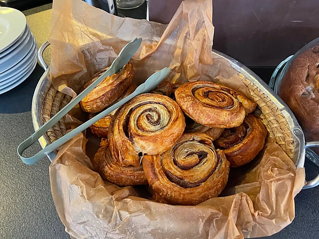 Korb mit frischen Zimtschnecken auf Backpapier, daneben eine Zange und gestapelte Teller