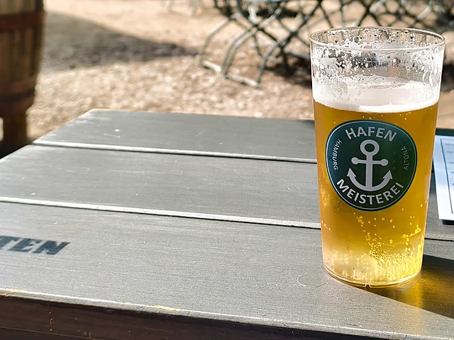 Glas Bier mit Hafenmeisterei-Logo auf einem grauen Holztisch im Biergarten, im Hintergrund Klappstühle im Schatten