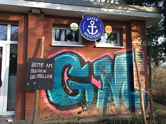 Backsteinkiosk mit blauem Hafenmeisterei-Schild, Graffiti-Wand und Kreidetafel „Bitte am Tresen bestellen“