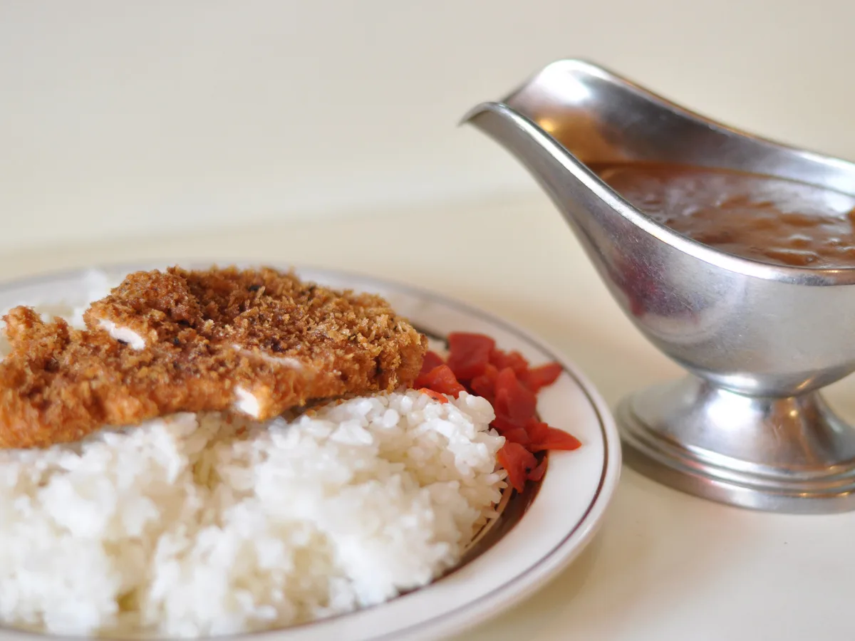 Paniertes Hähnchenschnitzel auf Reis, daneben eine Sauciere mit japanischem Curry
