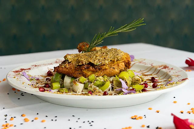 Kräuterkruste auf gebratenem Fisch mit Lauch, Granatapfelkernen und Rosmarin auf weißem Teller, drumherum Linsen und Blüten