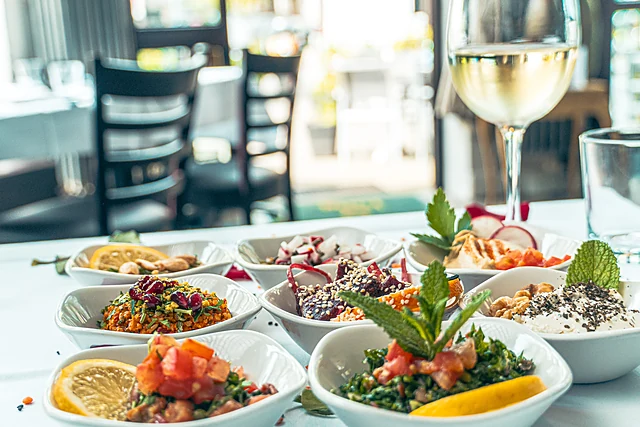 Kleine weiße Schälchen mit Taboulé, Salaten, Hummus und Dips vor dem Fenster, dazu ein Glas Weißwein