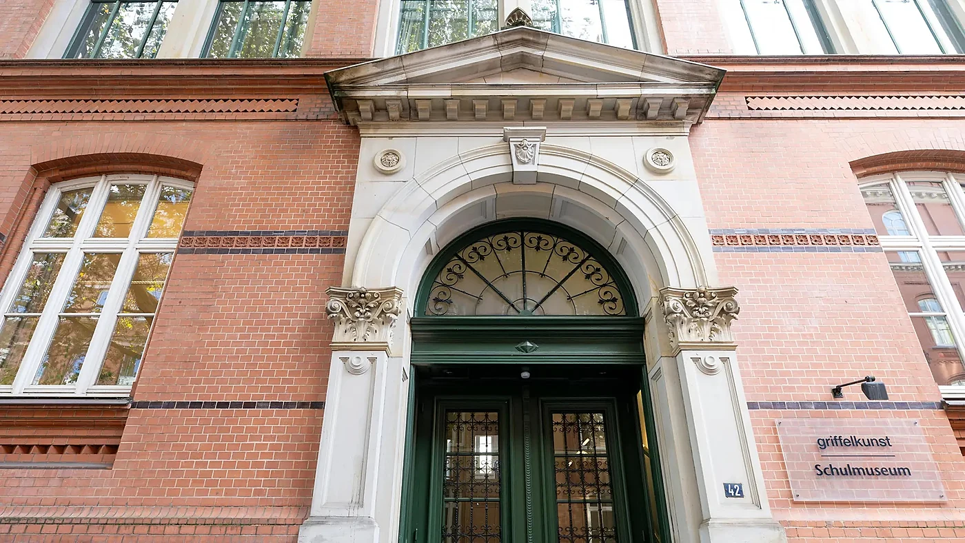 Sandsteinportal mit Rundbogen und grüner Holztür an der roten Backsteinfassade des Schulmuseums, daneben das Schild „Sch