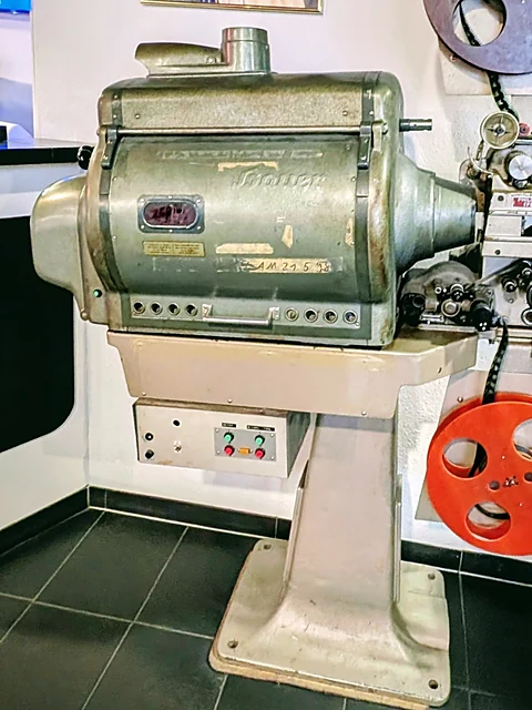 Alter Filmprojektor aus Metall auf einem Sockel, daneben eine rote Filmspule an der Wand