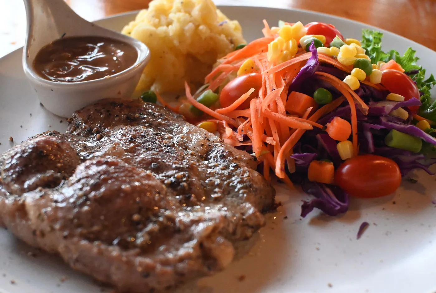 Gegrilltes Steak mit Kartoffelpüree, buntem Salat und einer Sauciere mit brauner Sauce auf einem weißen Teller