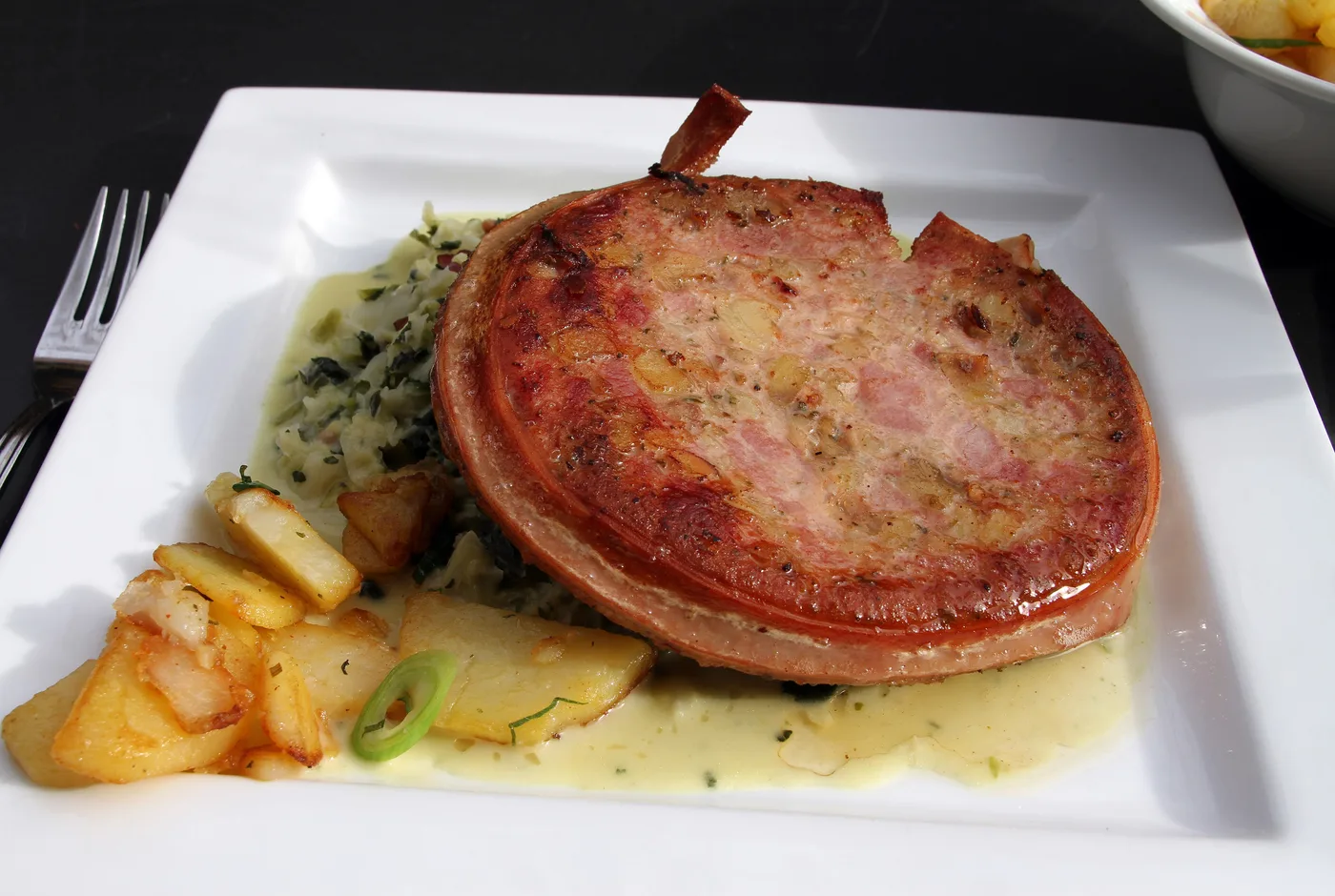 Scheibe Saumagen mit Bratkartoffeln, Kraut und heller Sauce auf einem weißen Teller