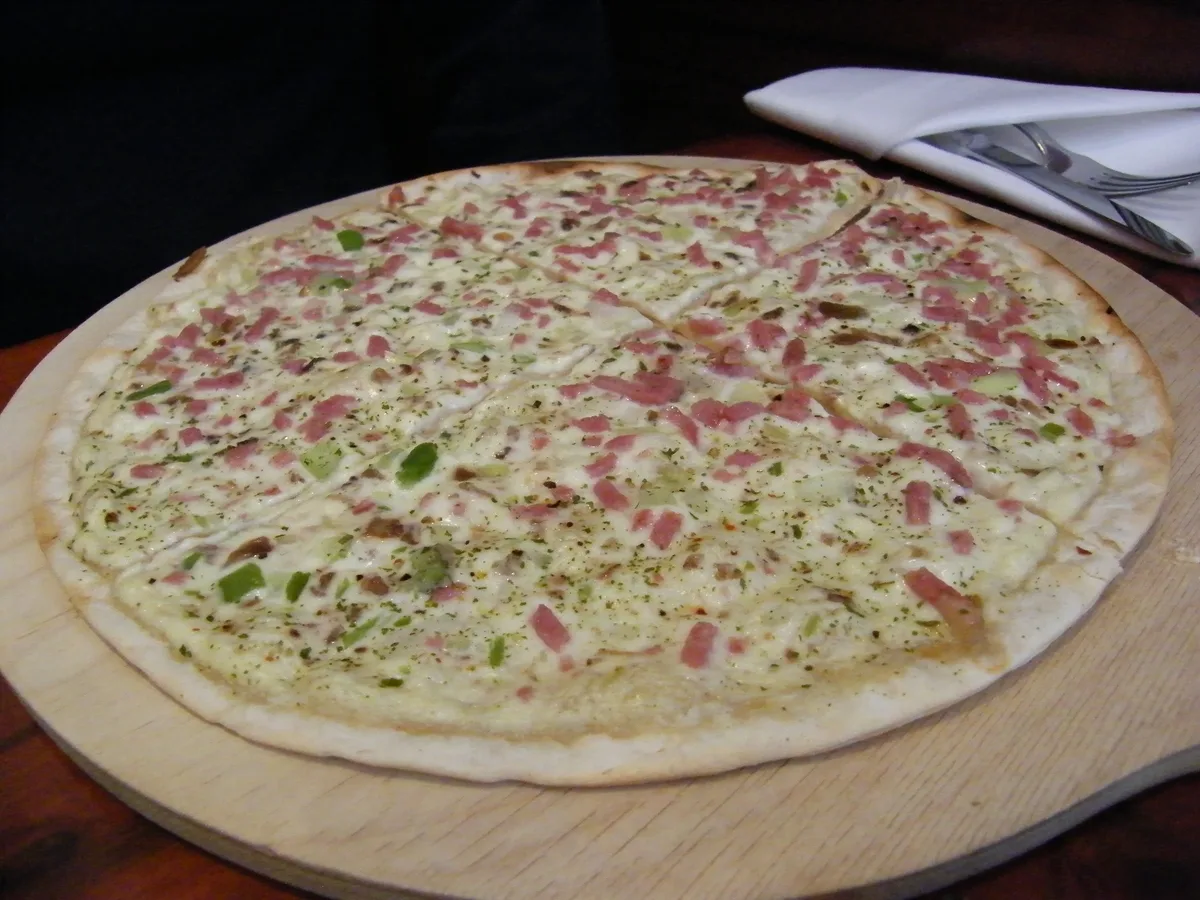 Flammkuchen mit Speckwürfeln, Zwiebeln und Creme auf einem runden Holzbrett