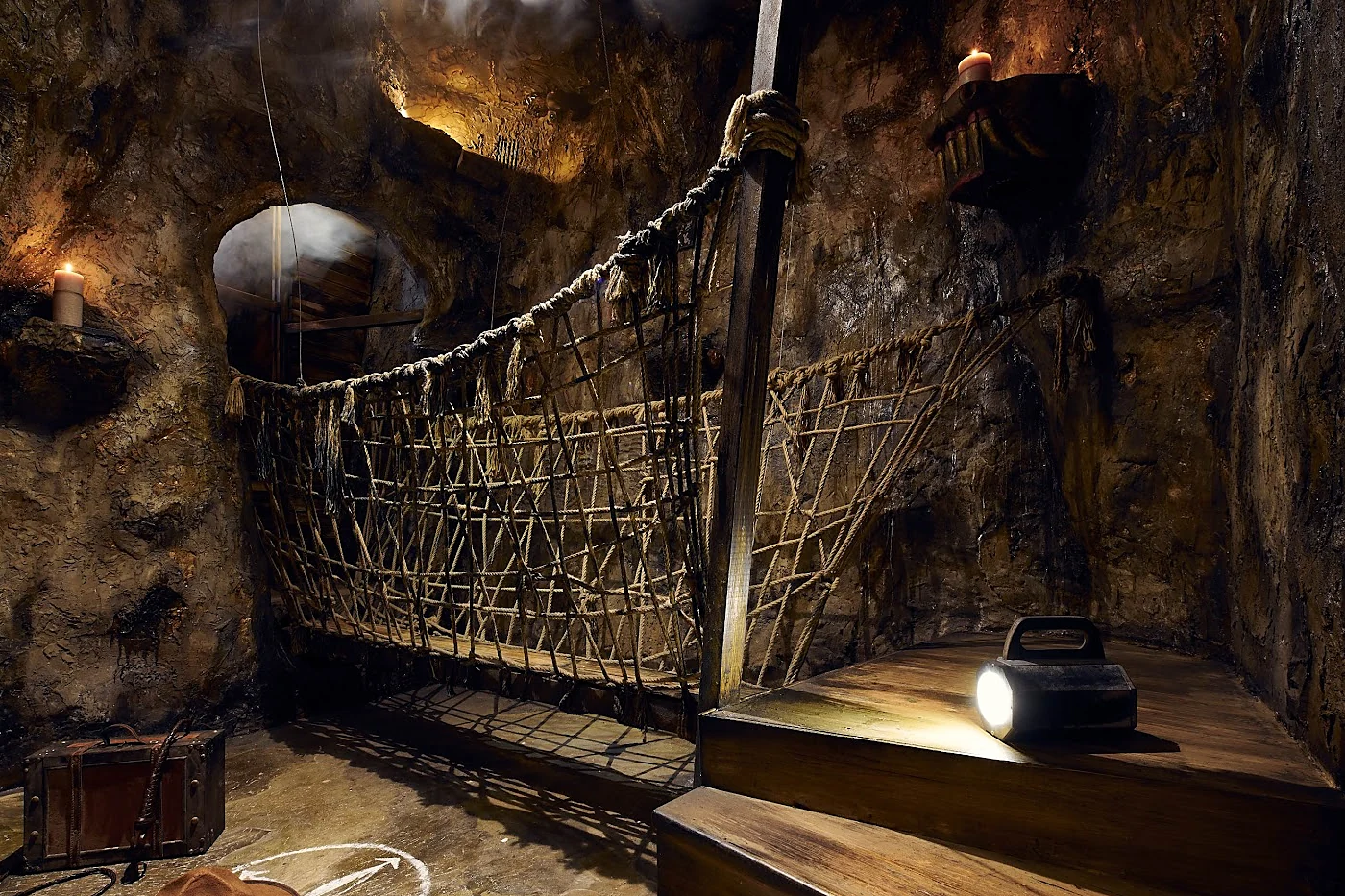 Escape-Room-Kulisse als Höhle mit Hängebrücke, Fackeln, altem Koffer und einer Taschenlampe auf einer Holzstufe