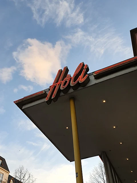 Roter Neonschriftzug Holi in Schreibschrift auf einem weit auskragenden Betonvordach vor blauem Himmel