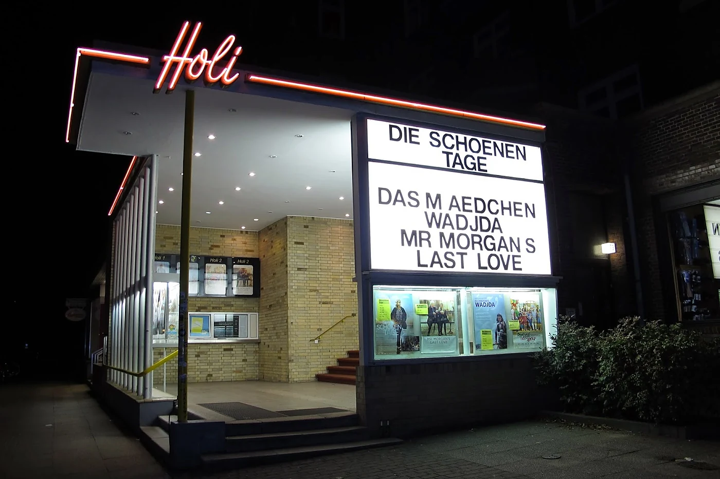 Kinoeingang bei Nacht mit rot leuchtendem Neonschriftzug, hell erleuchtetem Schaukasten und Programmtafel