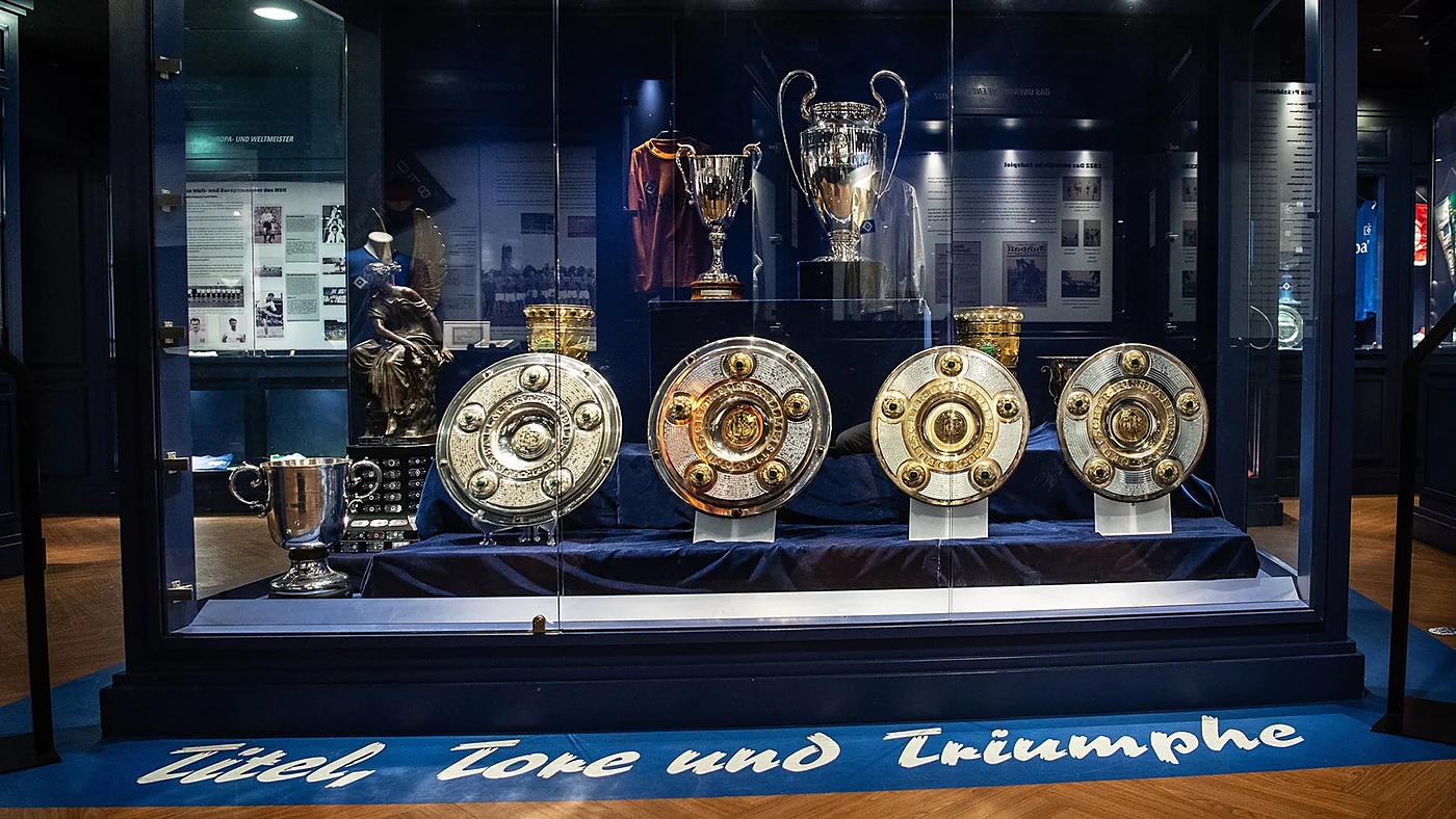 Beleuchtete Vitrine mit vier Meisterschalen, dem Europapokal der Landesmeister und weiteren Pokalen, davor der Schriftzu