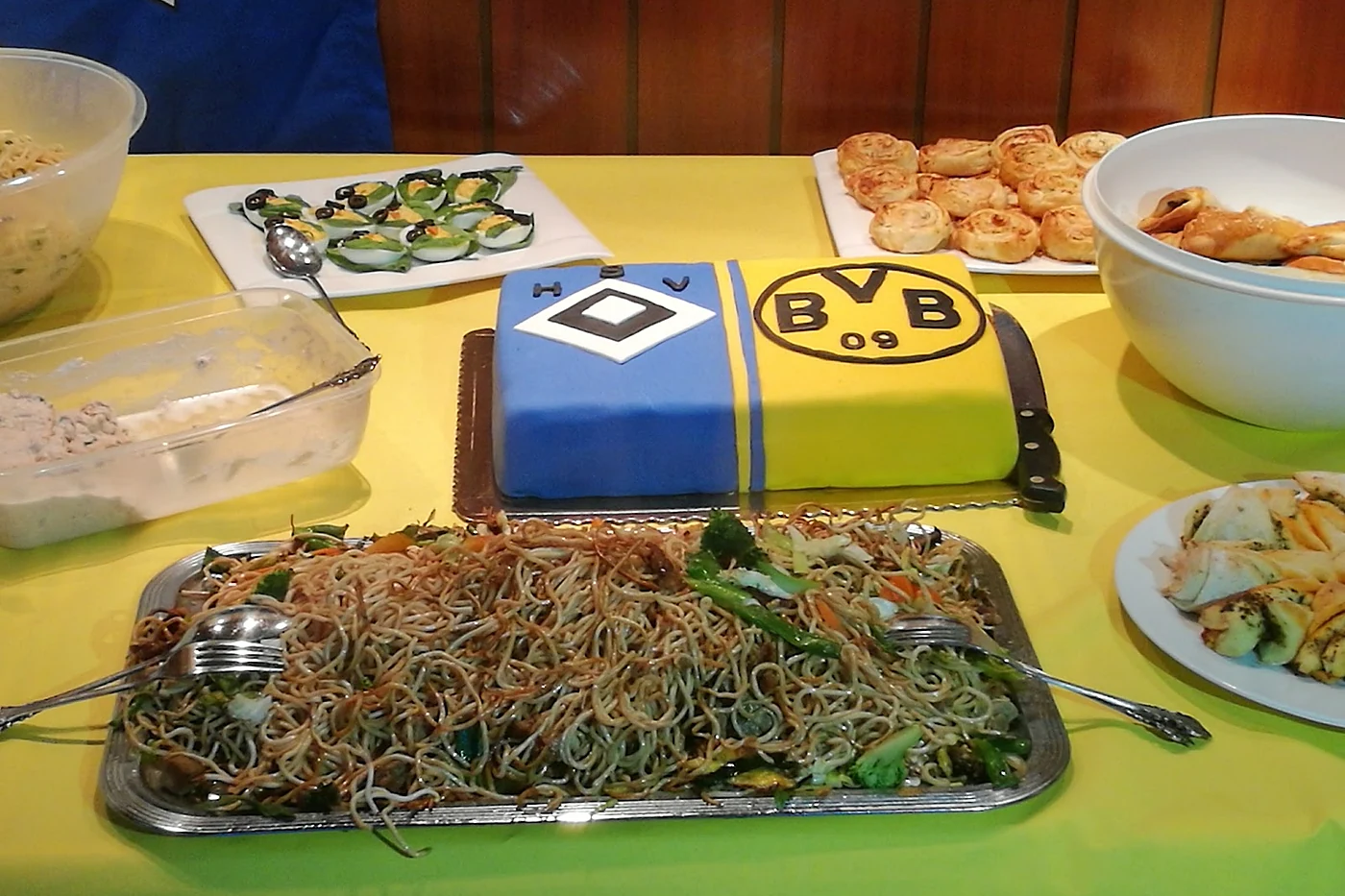 Buffet auf gelber Tischdecke mit Bratnudeln, Häppchen und einer Torte in HSV- und BVB-Farben