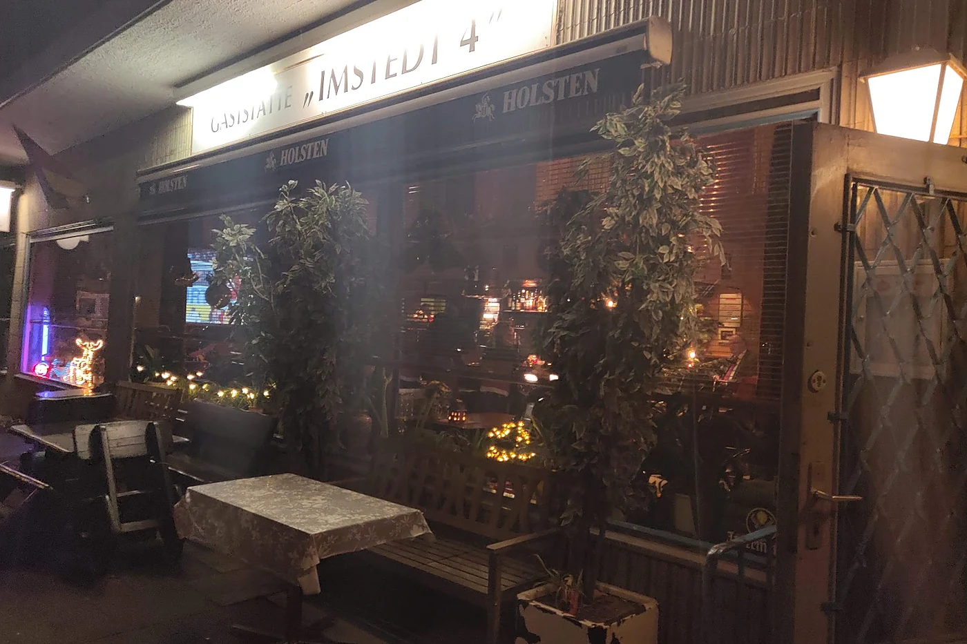 Die Gaststätte Imstedt 4 bei Nacht mit beleuchtetem Schild, Lichterketten und Pflanzen vor dem Fenster