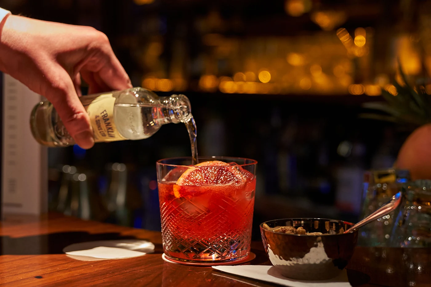 Barkeeper gießt Tonic aus einer kleinen Flasche in einen roten Cocktail mit Blutorangenscheibe auf dem Tresen